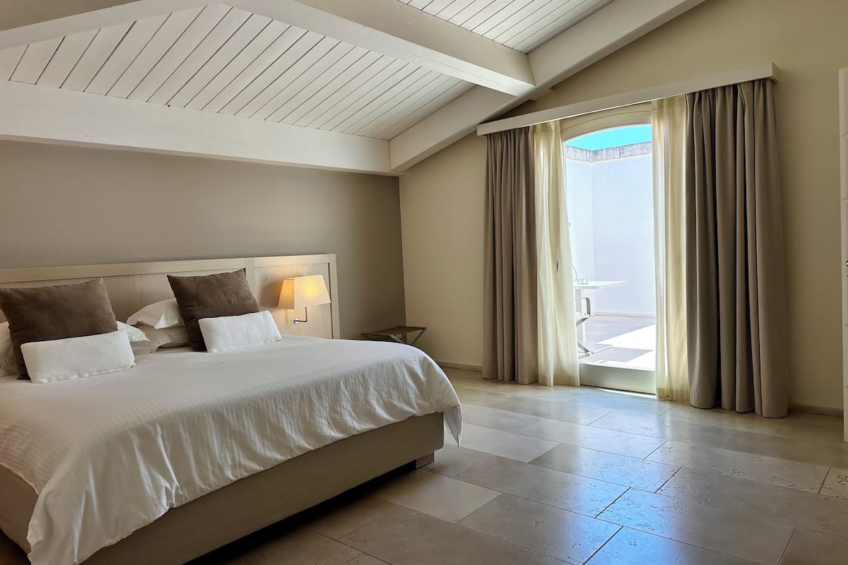 Habitación de hotel con una cama grande, una mesita de noche, una lámpara y vistas al exterior a través de un ventanal corredero.