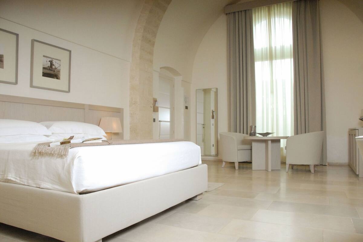 Una habitación de hotel con una cama grande, un escritorio, una silla, un ventanal con cortinas y cuadros en la pared.