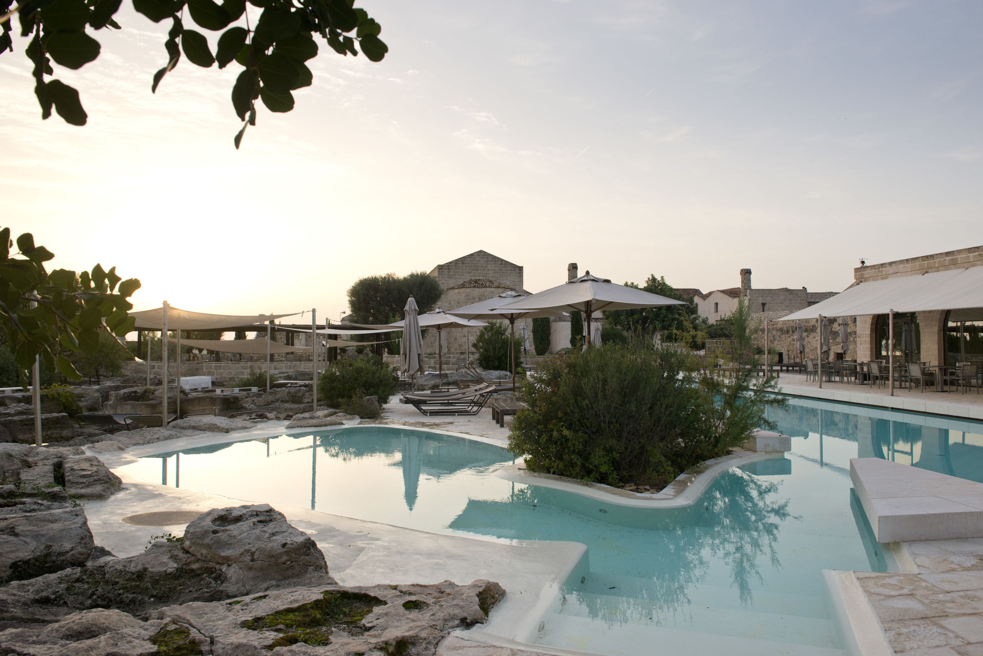 Un espace piscine doté de chaises longues et de parasols, entouré de bâtiments en pierre et de verdure.