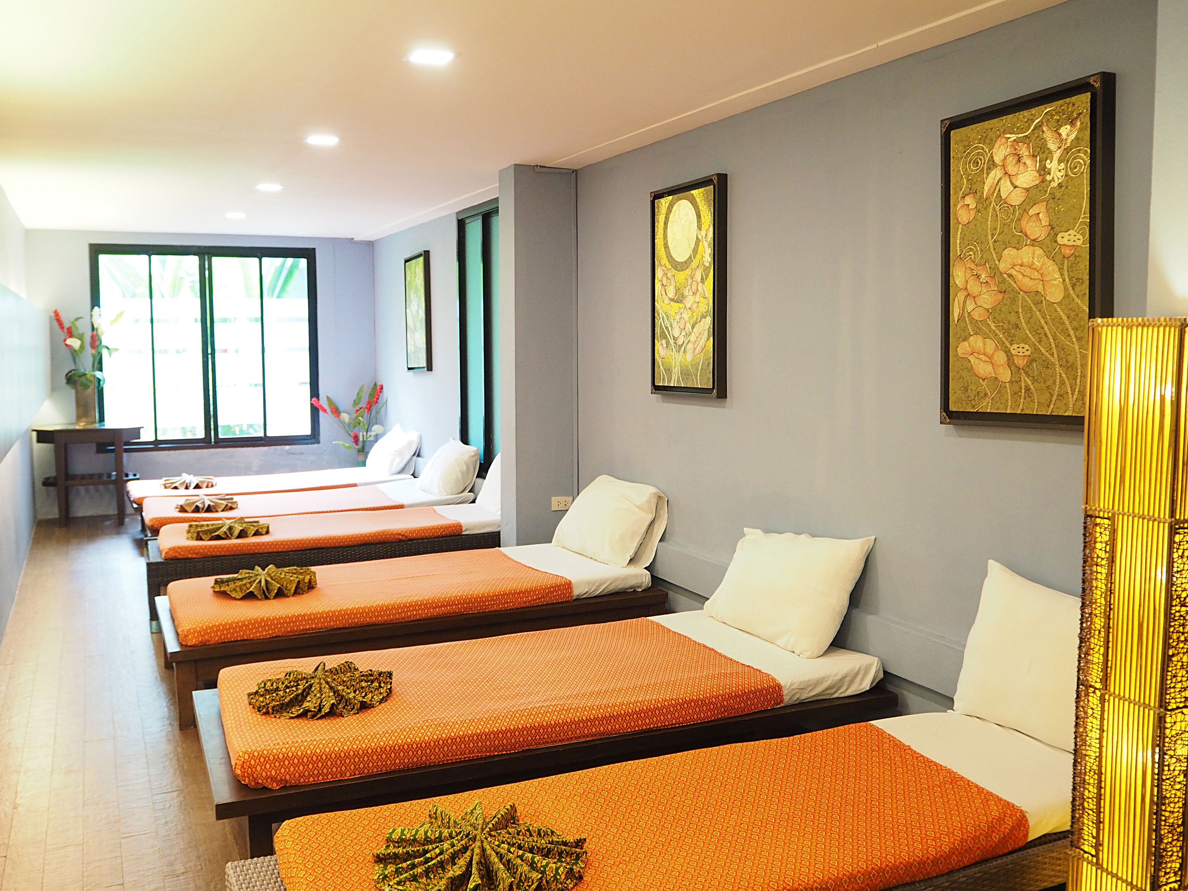 Ein Hotelzimmer mit vier Betten, jedes mit einer orangefarbenen Bettdecke und weißen Kissen. Zwei Fenster mit Vorhängen, ein Wandbild und ein kleiner Tisch mit einem Blumenstrauß.
