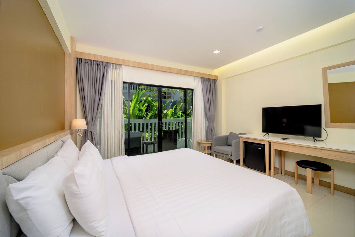Habitación de hotel con cama, escritorio, televisión y balcón con vista a follaje.