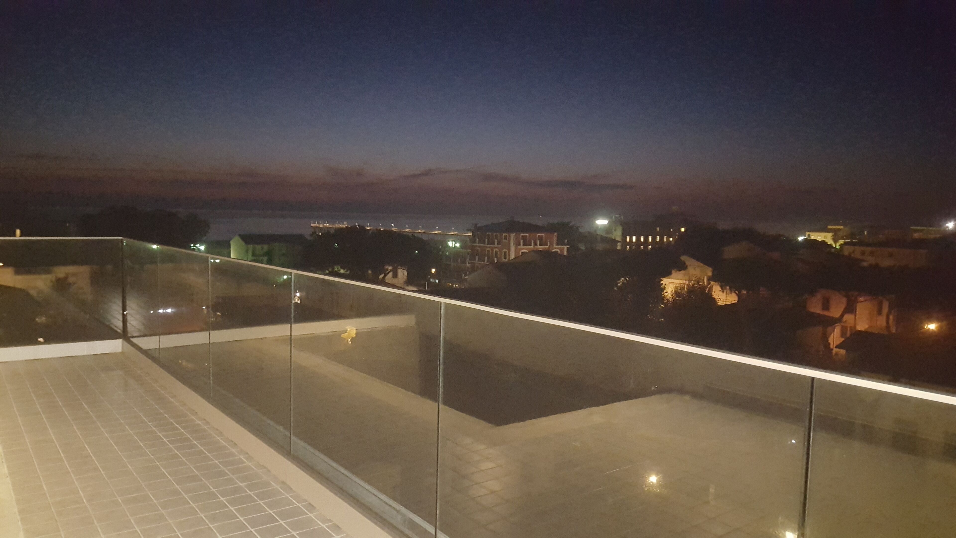 Vista nocturna desde un balcón, cielo despejado, edificios lejanos y suelo de baldosas.