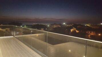 Vista nocturna desde un balcón, cielo despejado, edificios lejanos y suelo de baldosas.