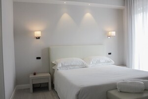 Una habitación de hotel con una cama grande, dos lámparas de noche, una mesita auxiliar pequeña y un ventanal con cortinas.