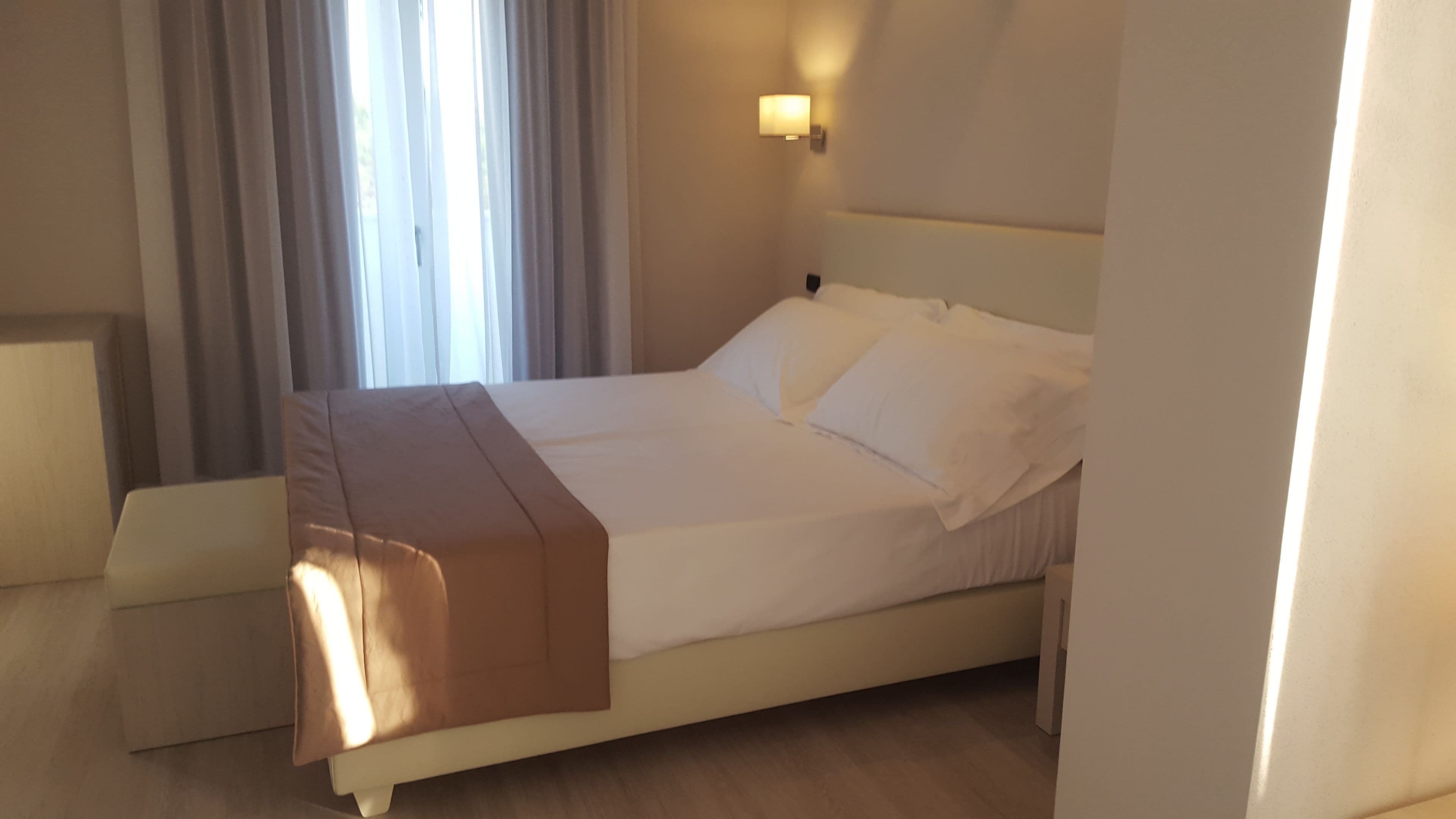 A neatly made bed with white linens and a brown throw blanket, a bedside table, and a wall-mounted light fixture.