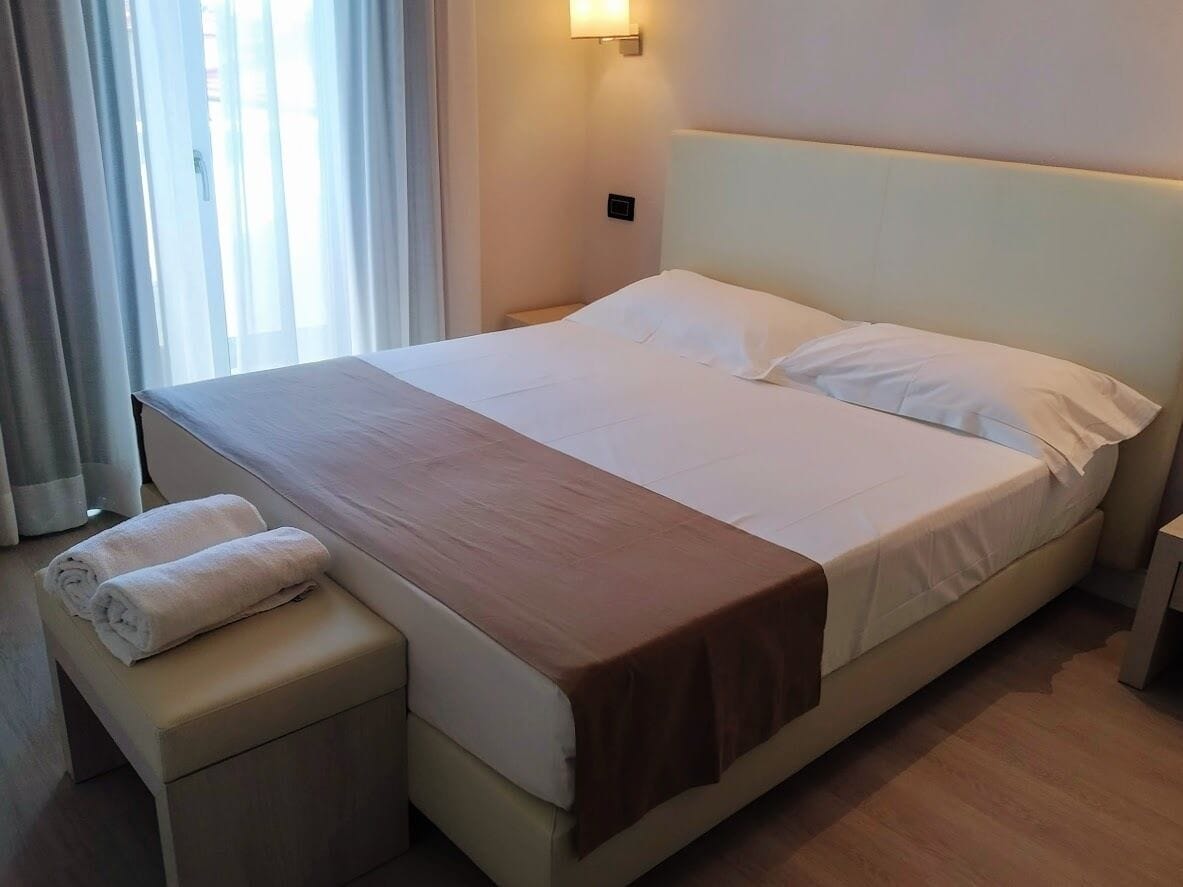 A large bed with a white headboard, a brown blanket, and two rolled towels on a matching ottoman.