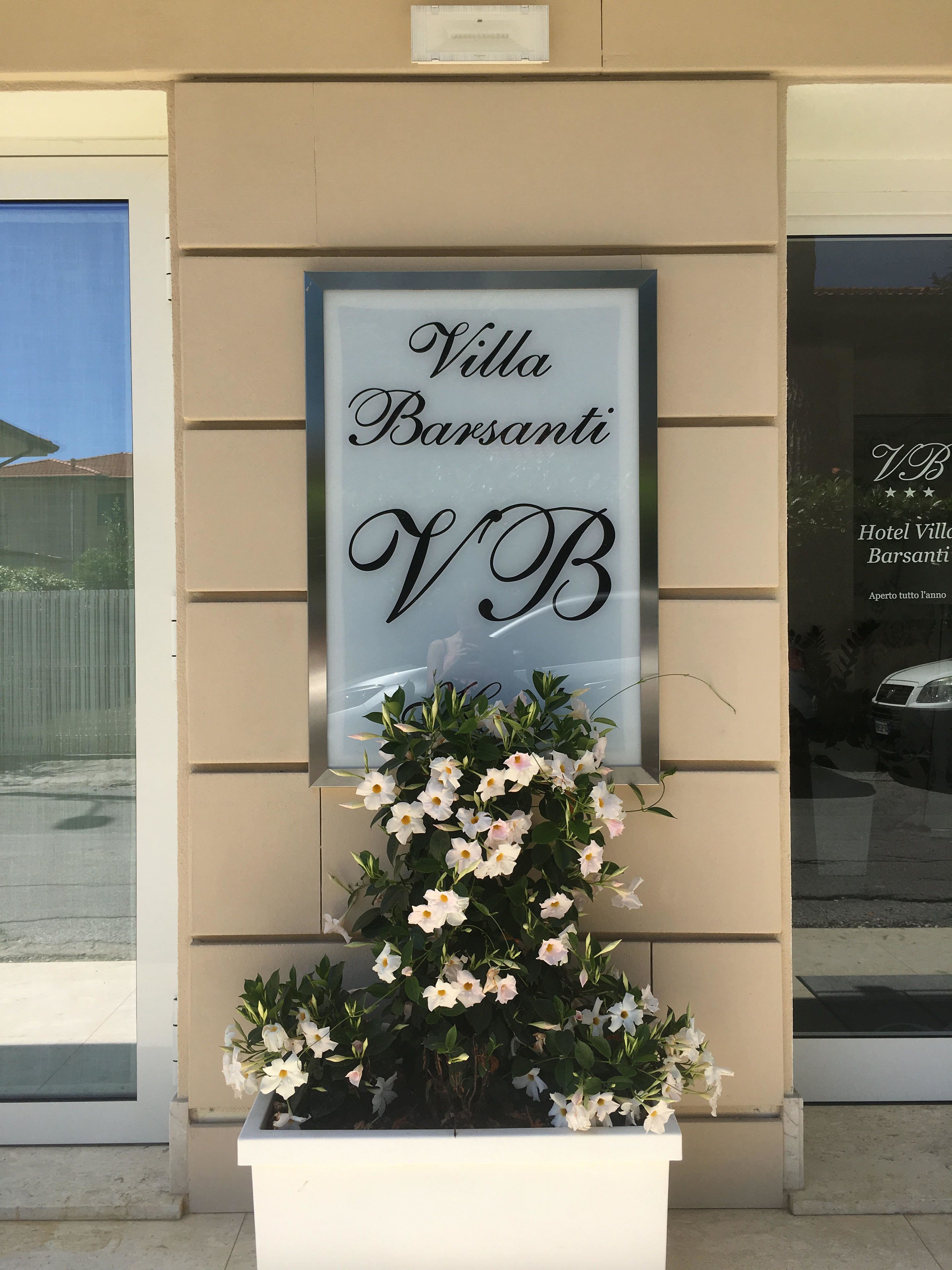 A sign for Villa Barsanti Hotel with a large 'VB' logo, a flower arrangement, and a reflection of a car in the window.
