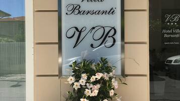 Placa do Hotel Villa Barsanti com um grande logotipo 'VB', um arranjo de flores e o reflexo de um carro na janela.