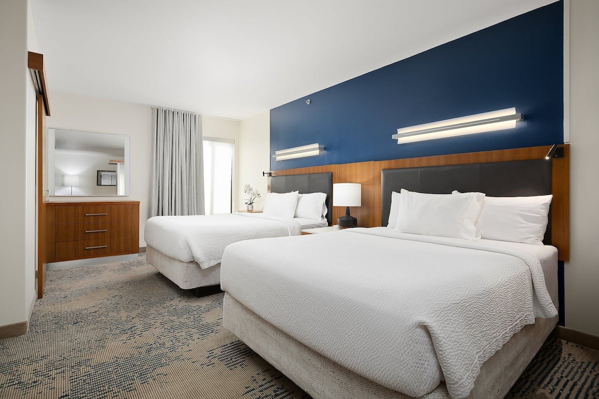 A hotel room with two beds, a wooden dresser, a mirror, and a large window with curtains.