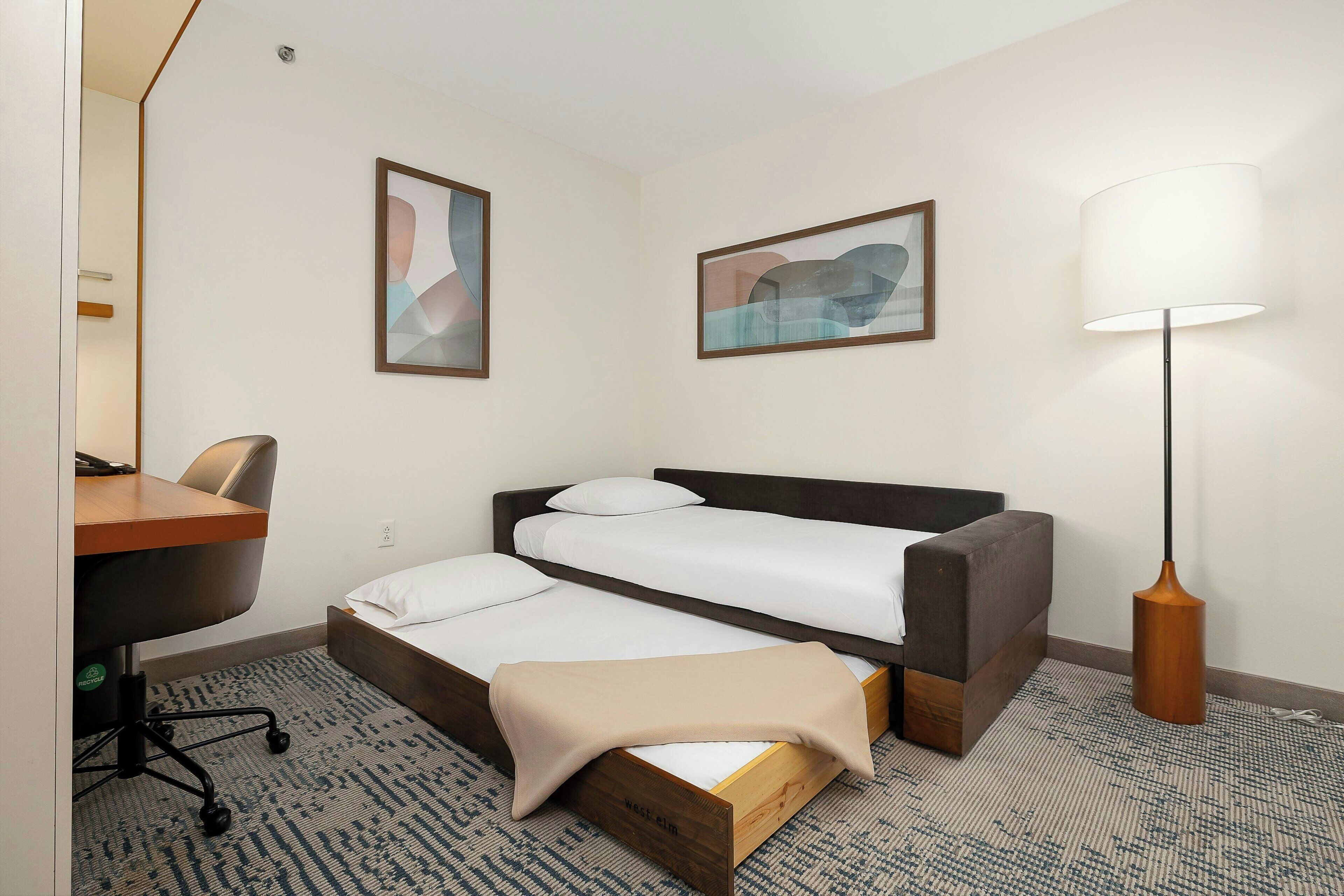 A hotel room with a bed, a desk, a chair, a lamp, and two framed pictures on the wall.