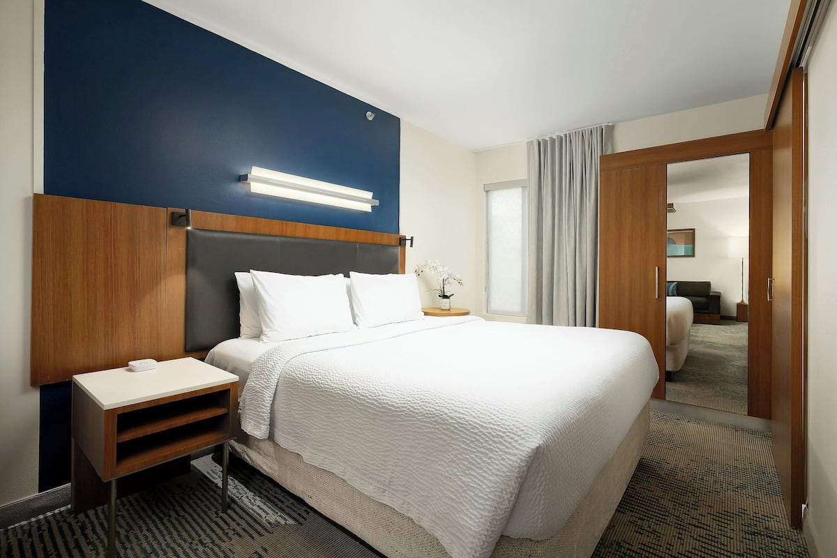 A hotel room with a large bed, a bedside table, a nightstand, a mirror, and a view of another room through an open door.