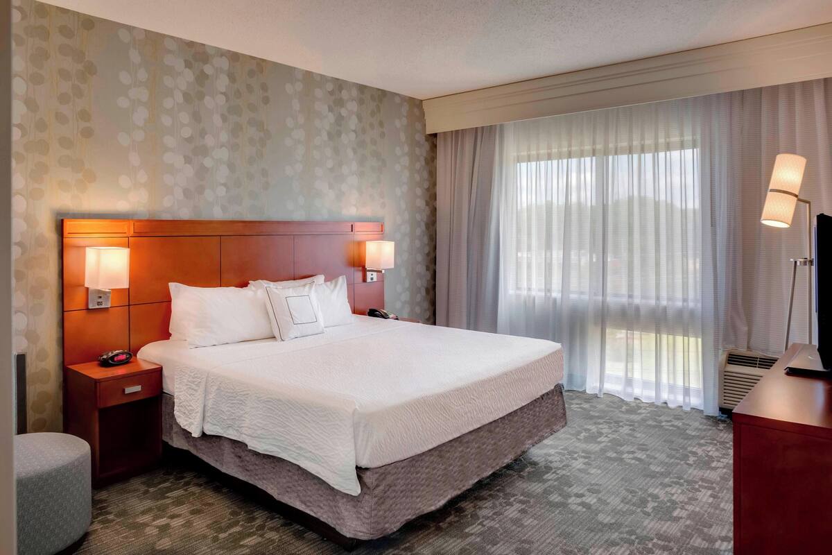 Ein Hotelzimmer mit einem großen Bett, zwei Nachttischlampen, einem Nachttisch, einem Fernseher und einem Fenster mit Vorhängen.