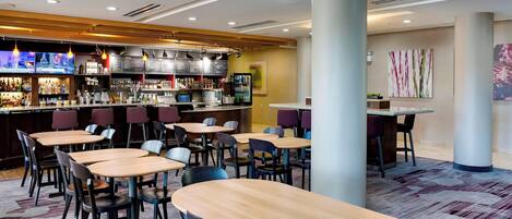 A modern restaurant with multiple tables and chairs, a bar area with a TV screen, and a menu board.