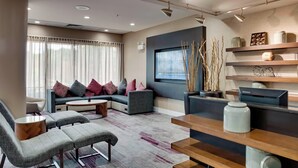 Un salon moderne avec un canapé d’angle, une table basse et un téléviseur. On y trouve des étagères ornées d’objets décoratifs et une fenêtre avec des rideaux.
