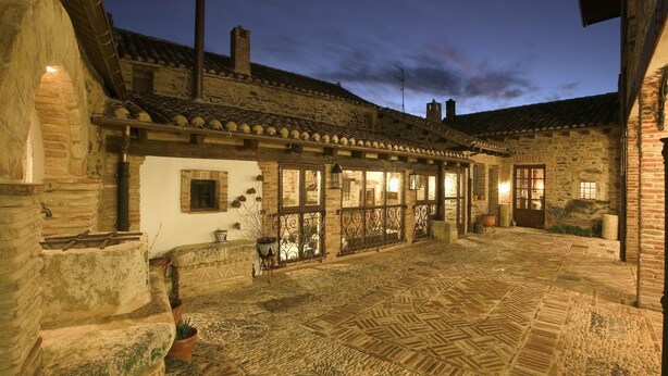 A stone courtyard with a tiled floor, a wooden pergola, and a rustic building with multiple windows and doors.
