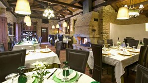 A restaurant with wooden beams, stone walls, and a fireplace. Tables are set with white tablecloths, green plates, and silverware.