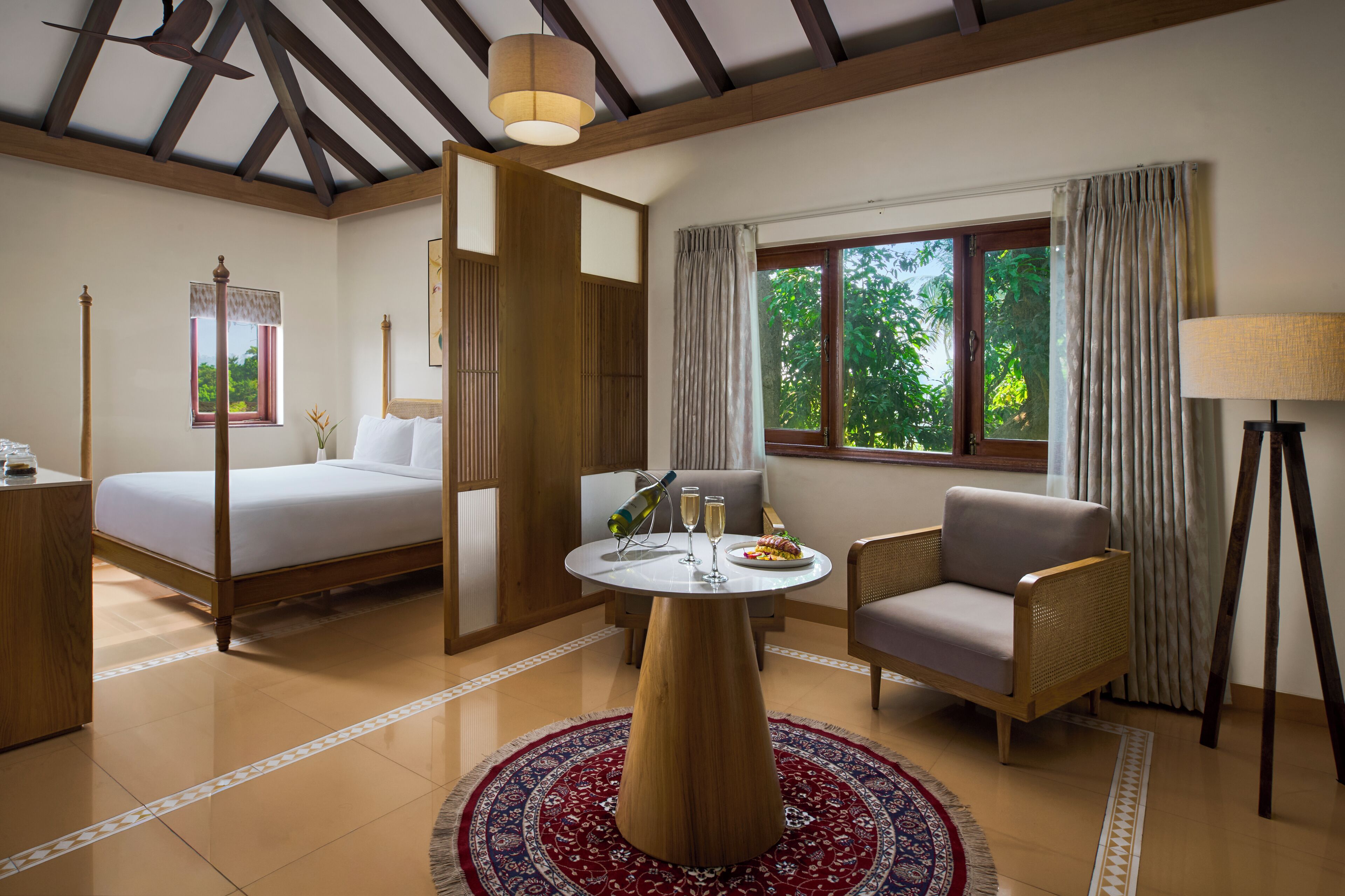A hotel room with a bed, a round table with two glasses and a plate of food, a chair, a lamp, and a window with curtains.
