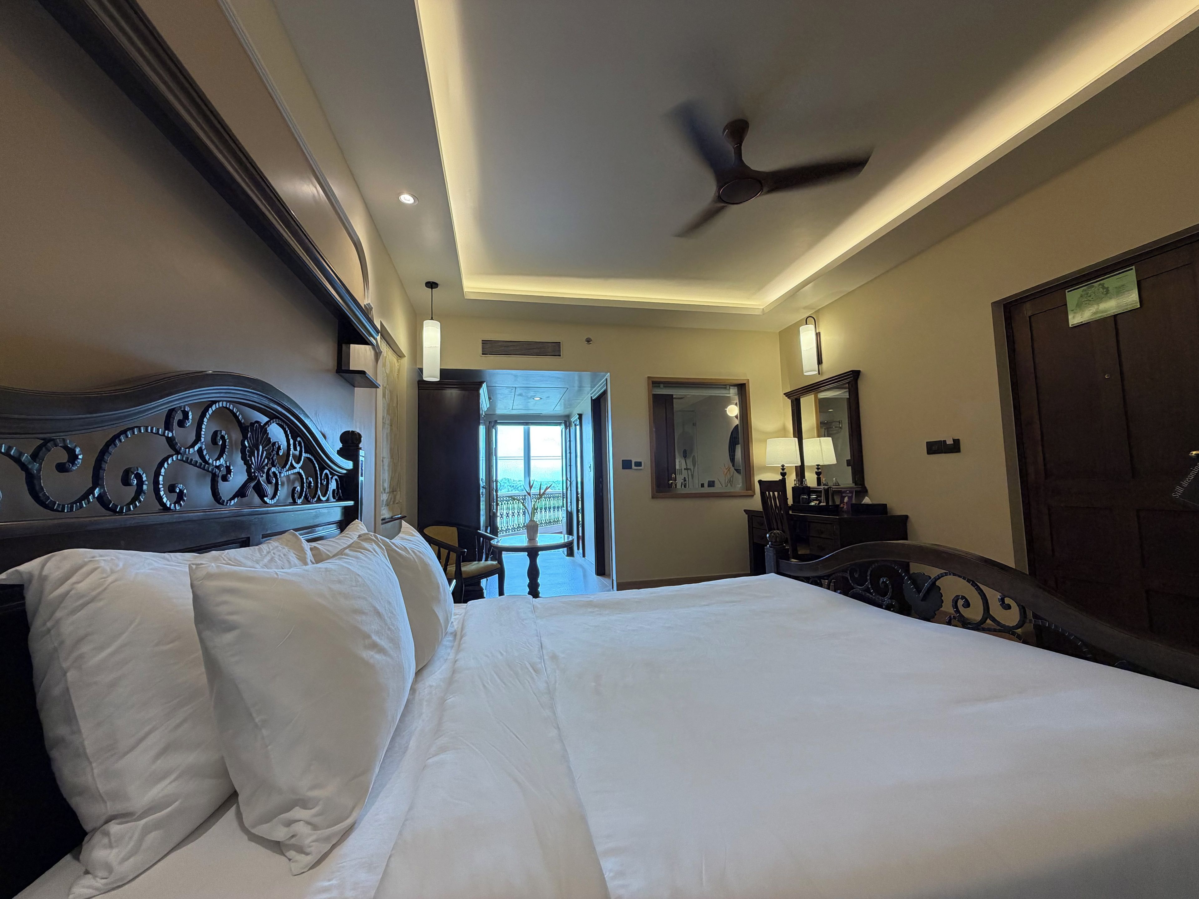 A hotel room with a large bed, a ceiling fan, a desk with a chair, a window with a view, and a door.