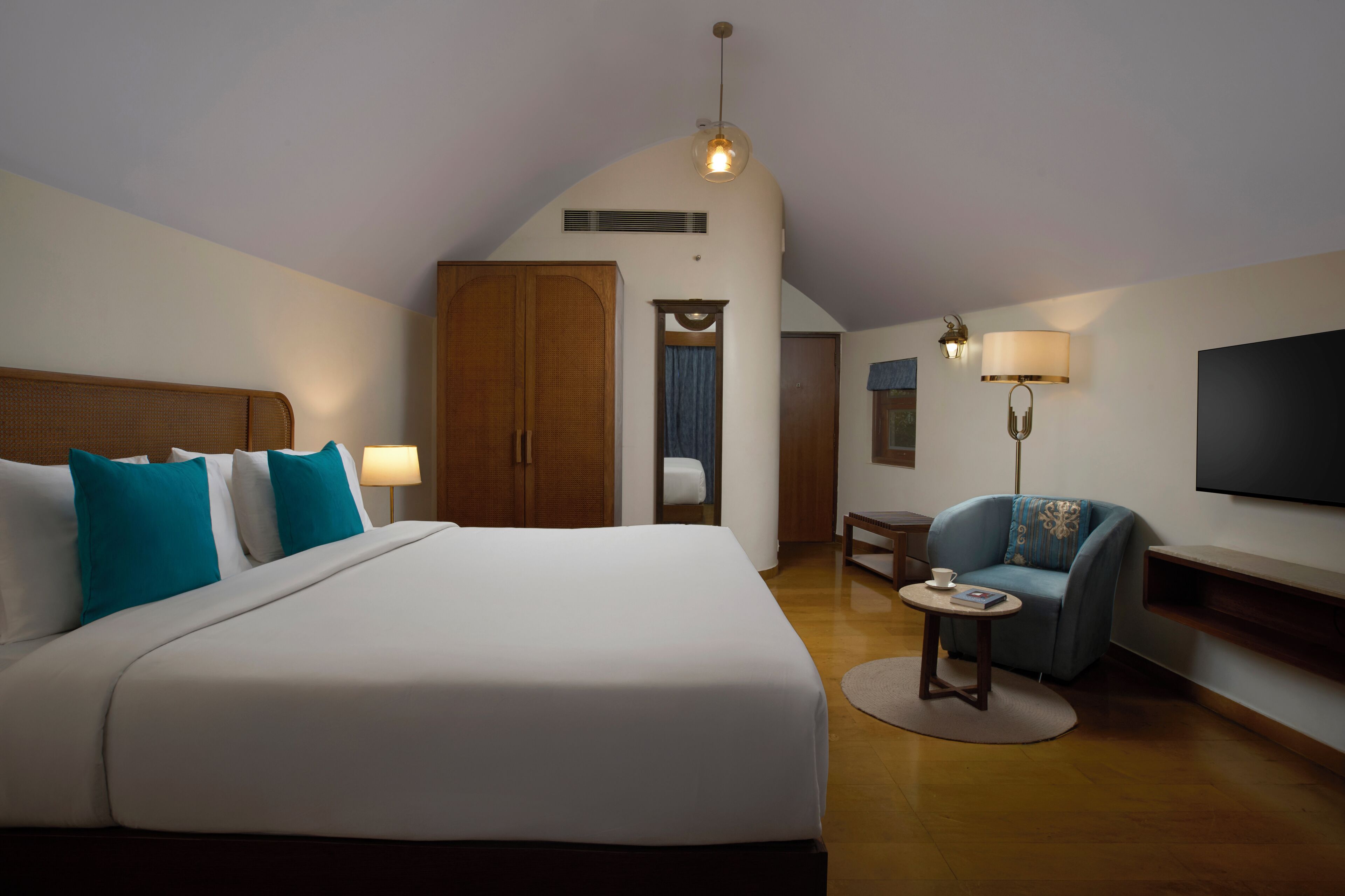 Una habitación de hotel con una cama grande, un armario de madera, una mesita pequeña, un sillón azul, un televisor y una lámpara en el techo.
