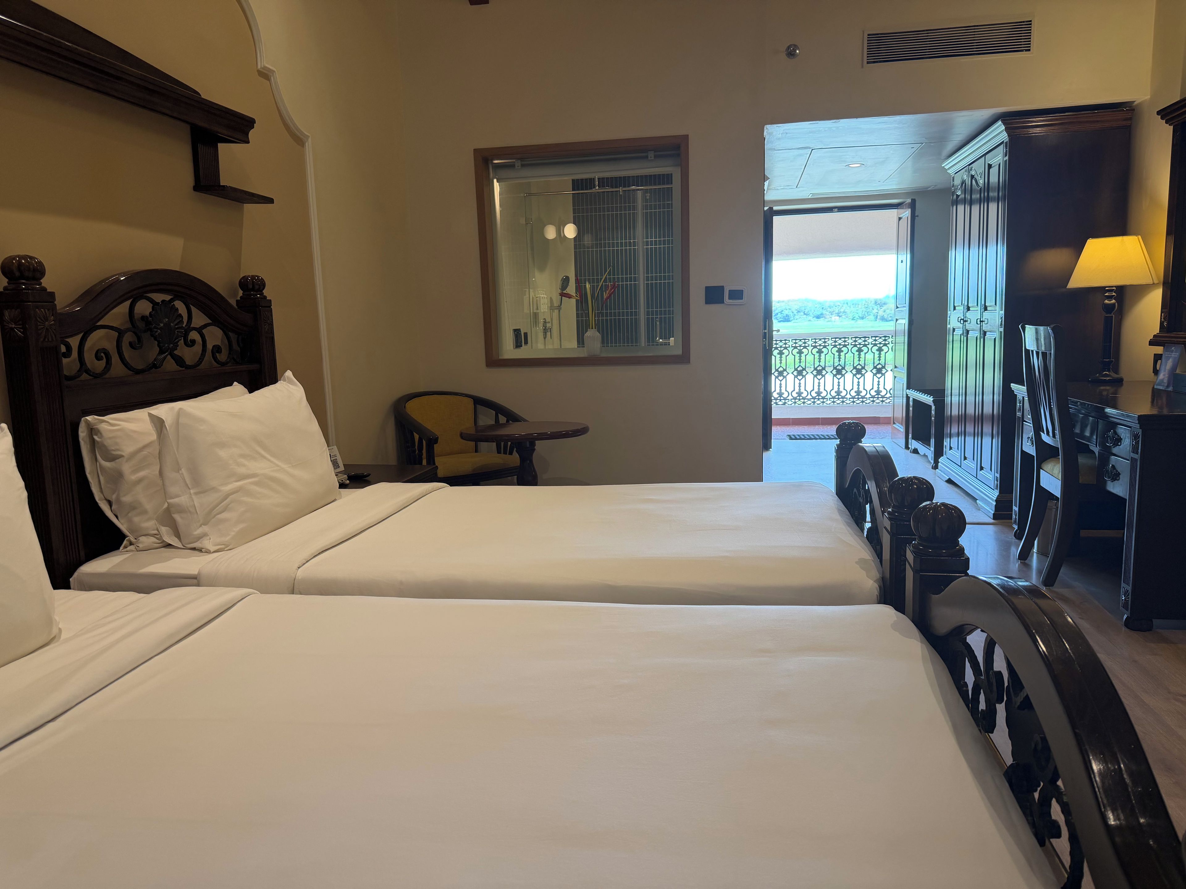 A hotel room with two beds, a chair, a small table, a lamp, and a view of the outdoors.