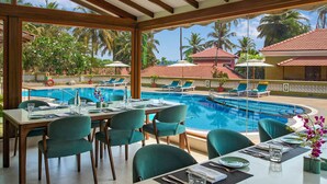 A poolside dining area with tables and chairs, a pool, and a view of palm trees and buildings. - Mercure Goa Devaaya Resort - An Accor Hotels Brand (Panaji)