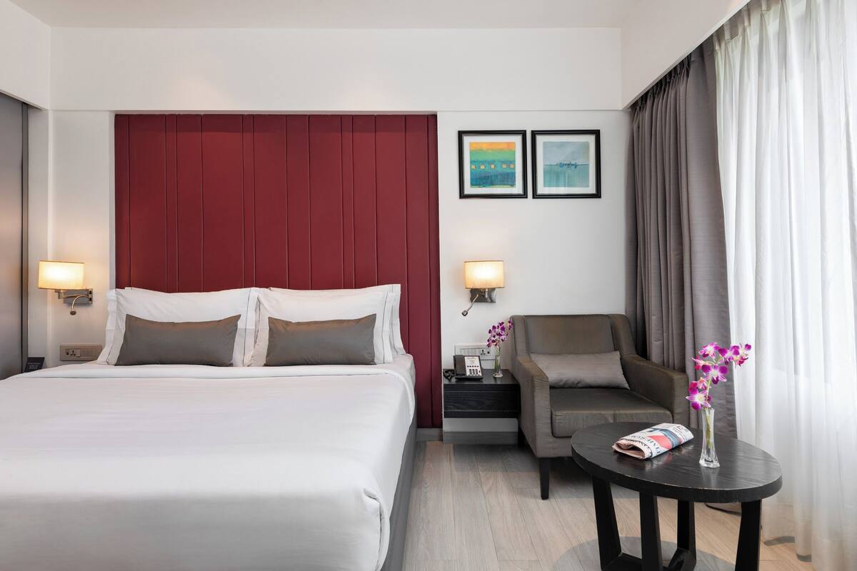 A hotel room with a large bed, a desk with a lamp, a chair, a small table with a vase of flowers, and two framed pictures on the wall.