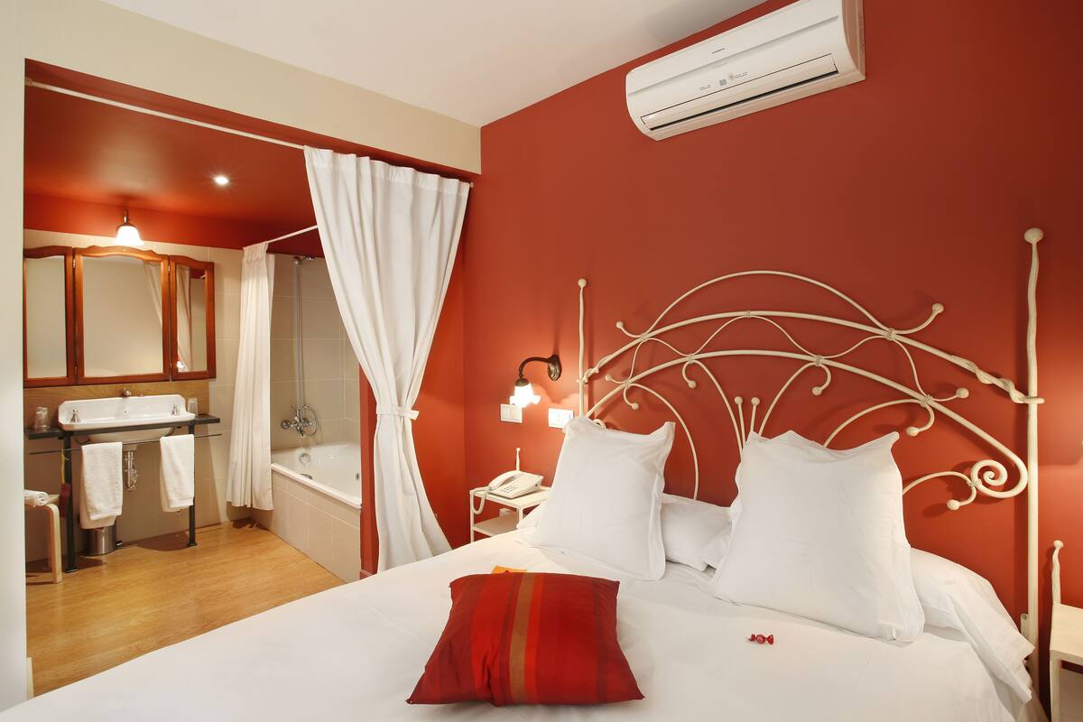 A hotel room with a red-walled bathroom, a white bed with a red pillow, and a white air conditioning unit.