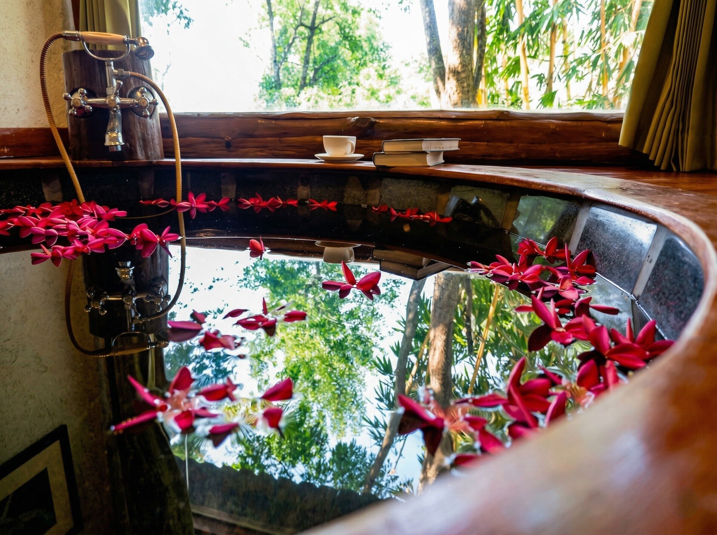 Una habitación con un espejo curvo grande que refleja árboles, un banco de madera, una mesita con libros y una fuente decorativa con flores rojas.
