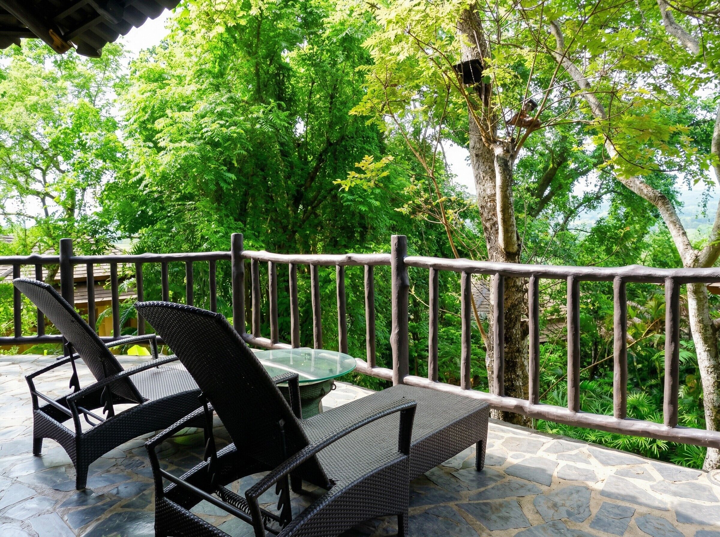 A patio with wicker furniture and a glass table, surrounded by a stone floor and a wooden railing.