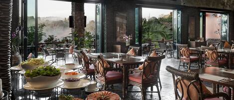 A restaurant with high ceilings, large windows, and a view of the outdoors. Tables are set with plates, bowls, and chairs.