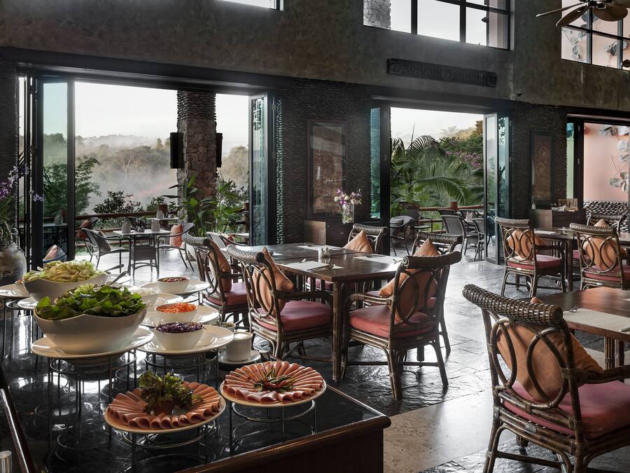Ein Restaurant mit hohen Decken, großen Fenstern und Blick ins Freie. Tische sind mit Tellern, Schüsseln und Stühlen gedeckt.
