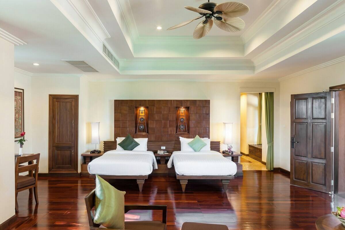 A hotel room with two beds, wooden flooring, a ceiling fan, and a wooden headboard with wall-mounted lights.