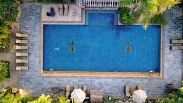 An aerial view of a rectangular swimming pool with a tiled blue interior, surrounded by lounge chairs and umbrellas, with a stone wall and tropical plants.