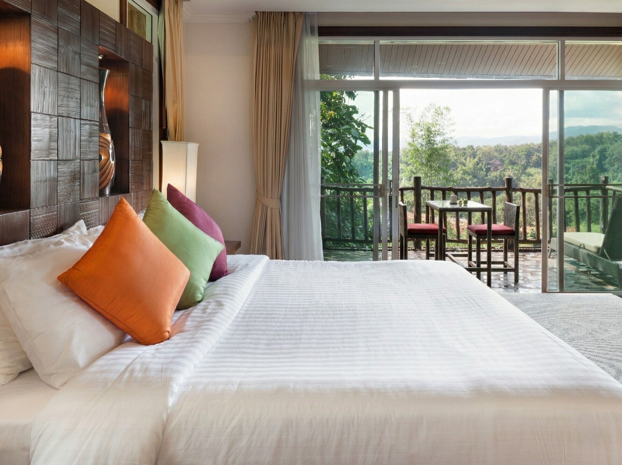 A hotel room with a large bed, a balcony with a table and chairs, and a view of the outdoors.