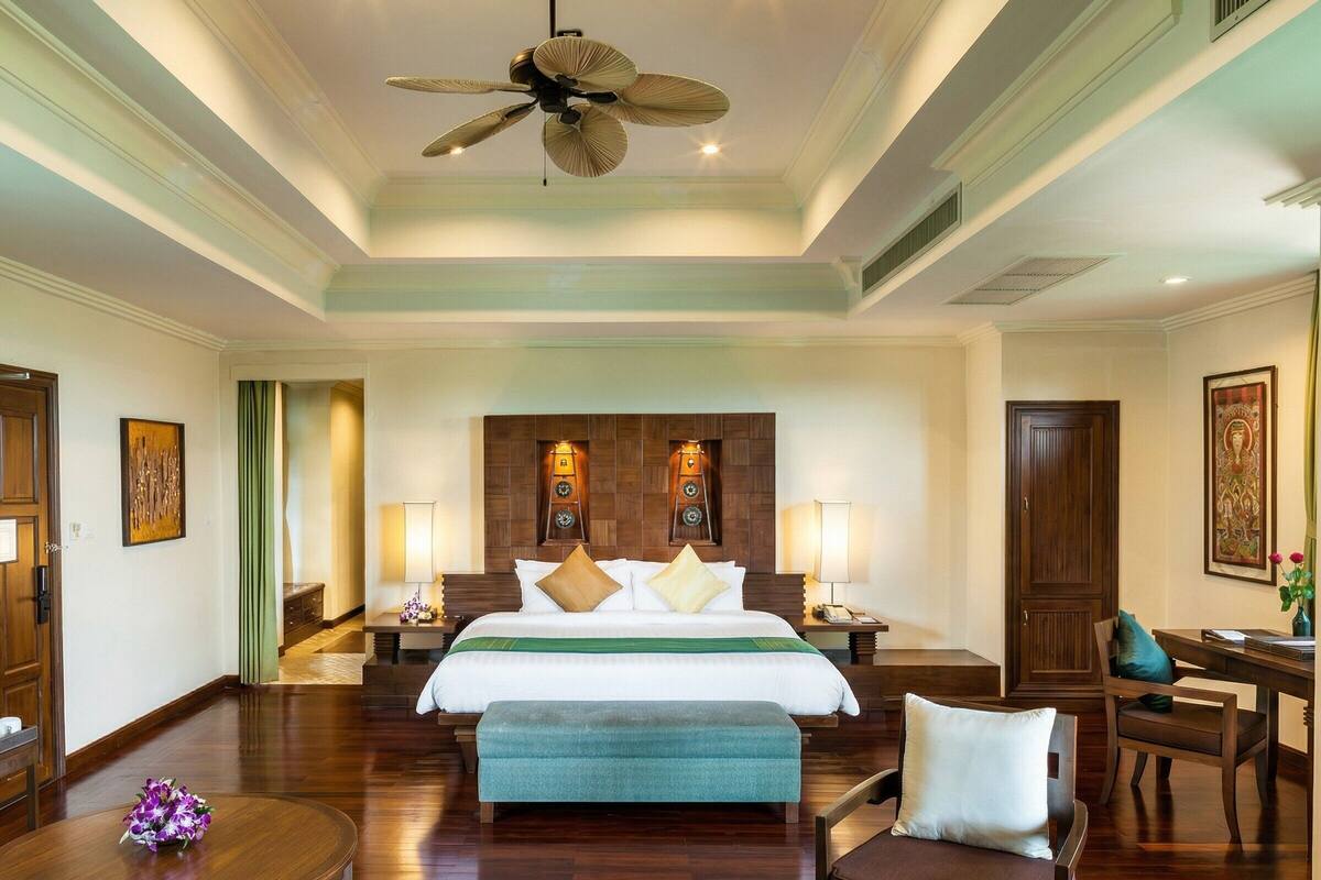A hotel room with a large bed, a ceiling fan, a desk with a chair, and a small table with a vase of flowers.