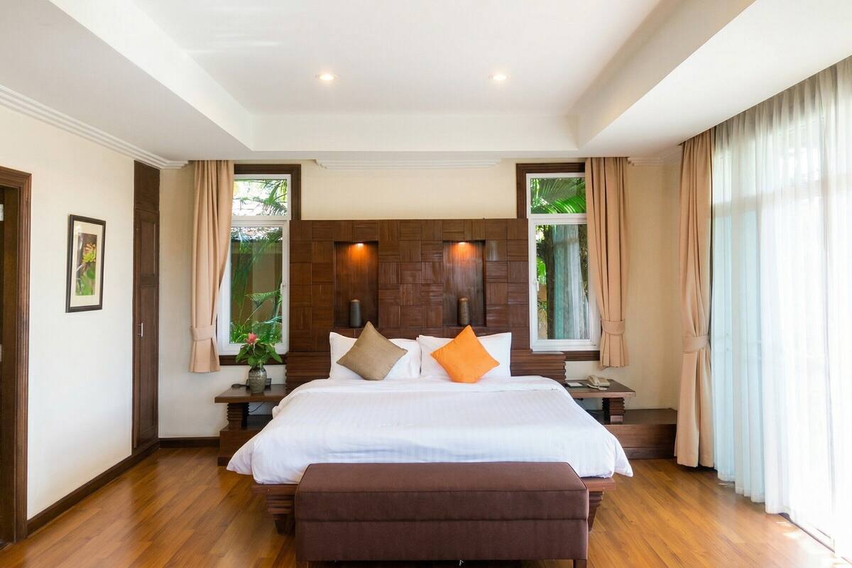 A hotel room with a large bed, wooden headboard, two bedside tables, a nightstand, a window with curtains, and a plant on the nightstand.