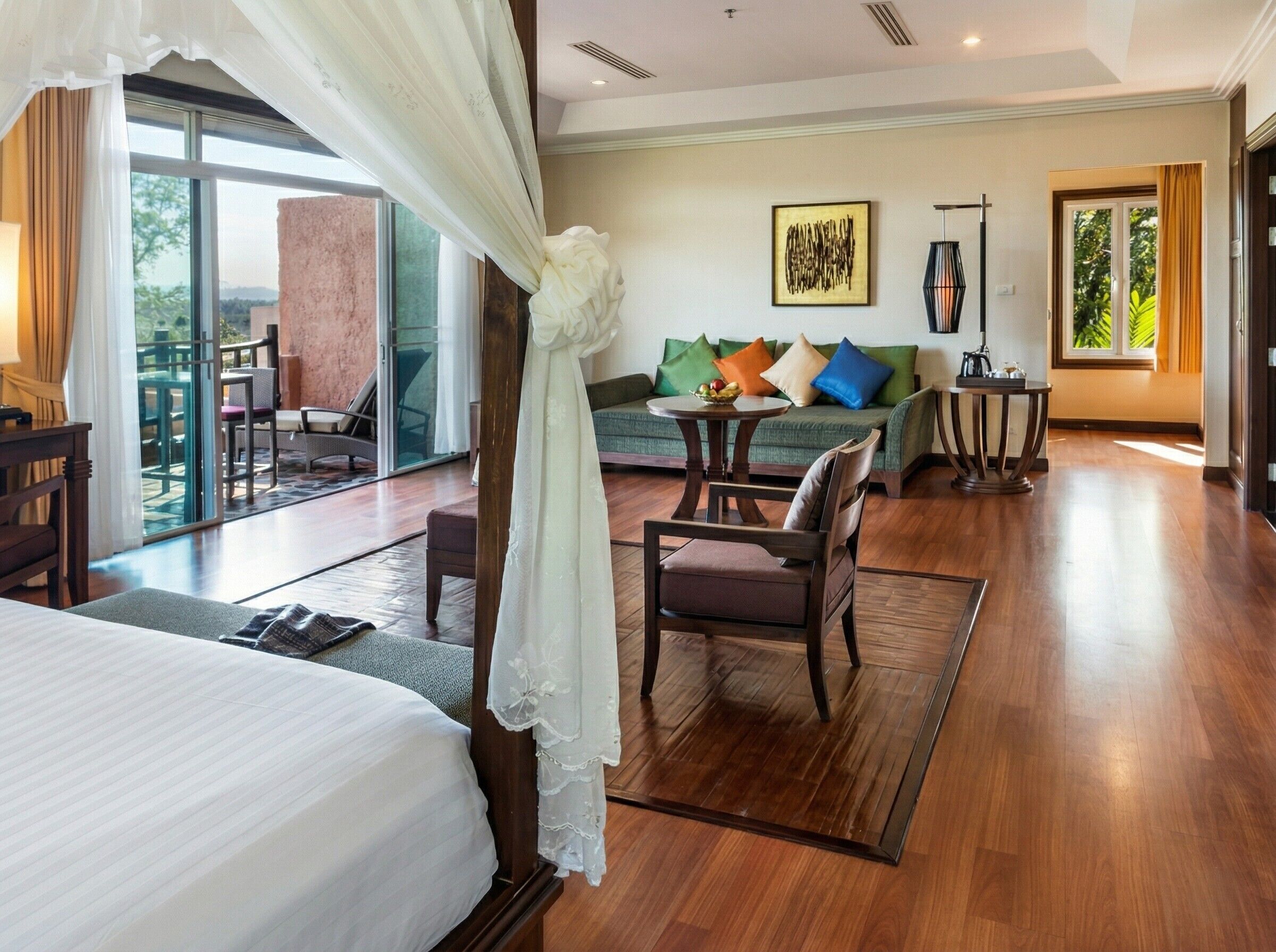 A hotel room with a bed, a sofa, a dining table, and a chair. There is a balcony with a view of the outdoors.