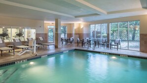 Ein Swimmingpool mit Liegestühlen und Tischen, großen Fenstern und Blick auf Bäume.