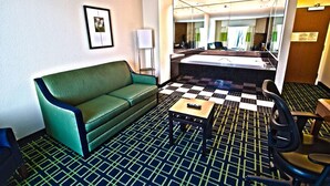 Habitación de hotel con sofá verde, una mesita, suelo a cuadros, un espejo grande y un baño con lavamanos y espejo.