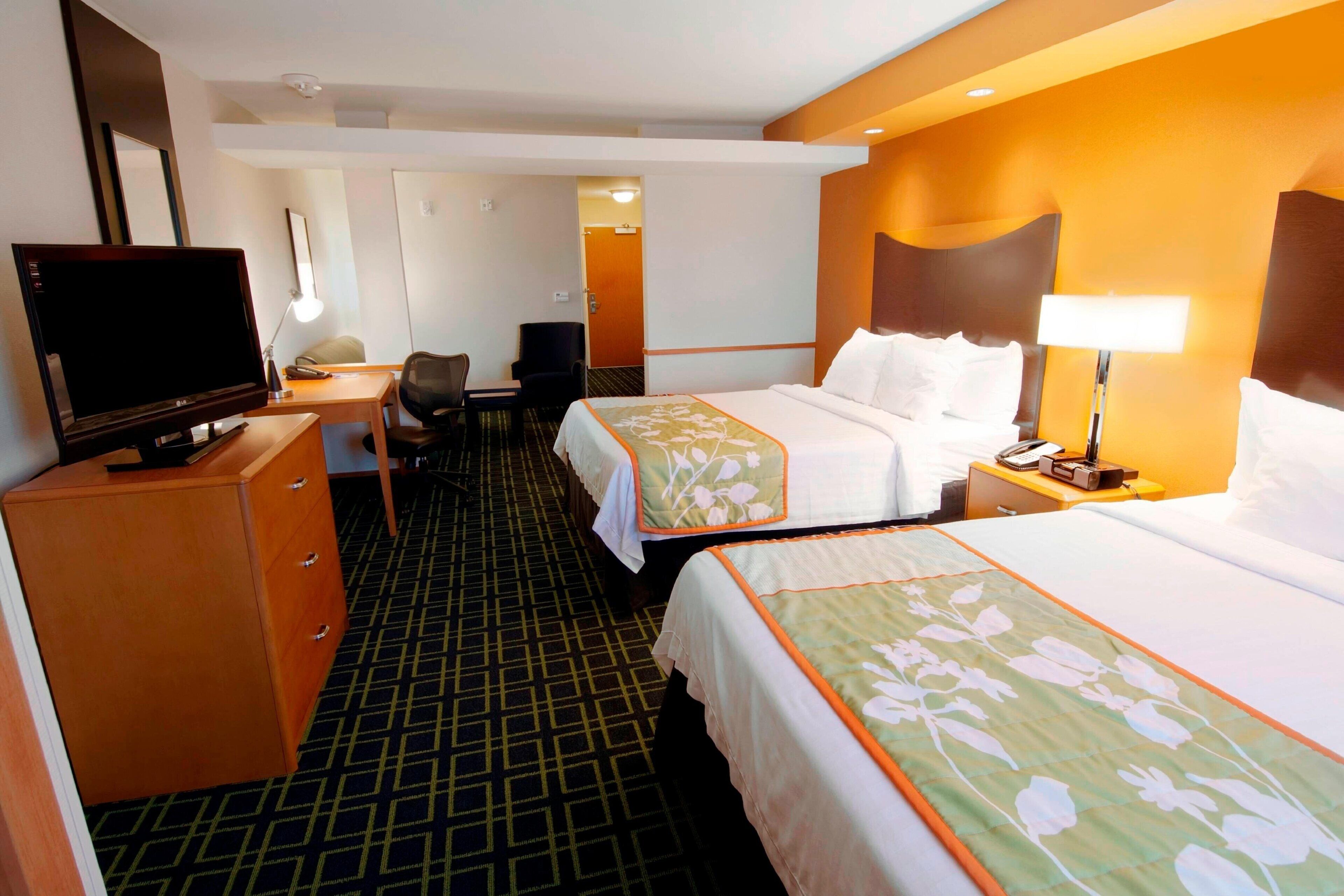 Habitación de hotel con dos camas, un escritorio, una silla, un televisor y una mesita de noche con lámpara.