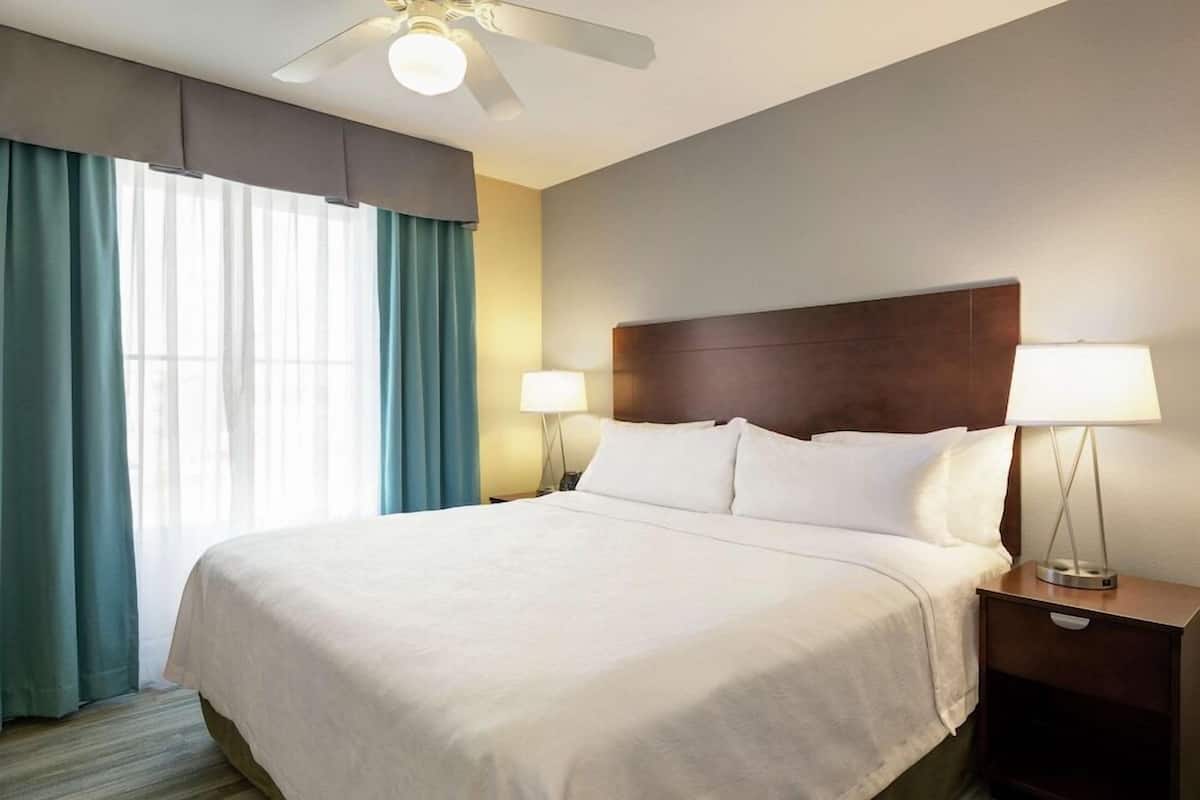 Habitación de hotel con una cama grande, dos lámparas de noche, un ventilador de techo y una ventana con cortinas.