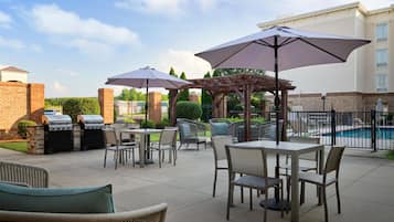Un patio exterior con mesas y sillas, una parrilla, sombrillas y una piscina al fondo.