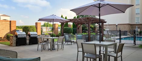 Un patio exterior con mesas y sillas, una parrilla, sombrillas y una piscina al fondo.