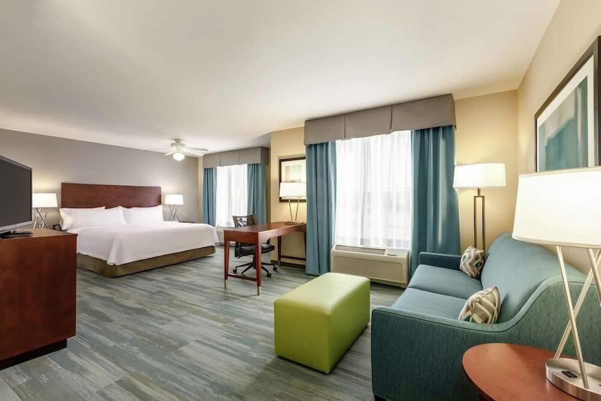 Una habitación de hotel con cama, escritorio, silla, sofá, reposapiés, televisor y lámpara.