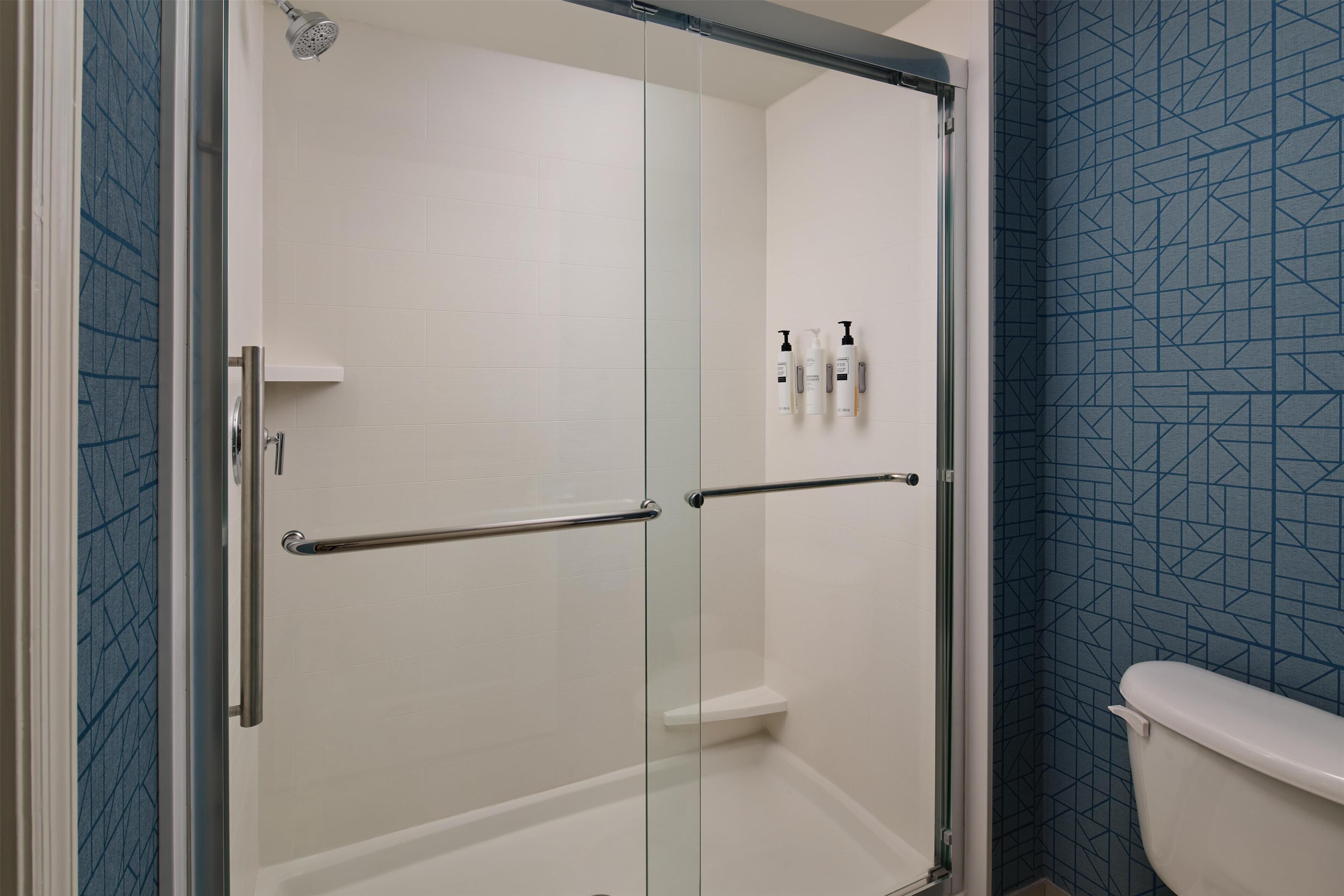 A glass-enclosed shower stall with a white tiled wall, a metal handrail, and a toilet with a blue patterned wall in the adjacent room.
