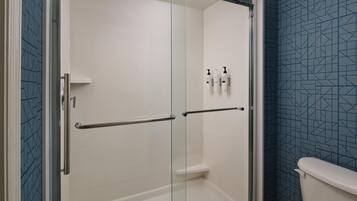 A glass-enclosed shower stall with a white tiled wall, a metal handrail, and a toilet with a blue patterned wall in the adjacent room.