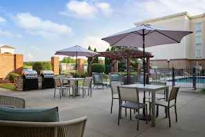 Un patio exterior con mesas y sillas, una parrilla, sombrillas y una piscina al fondo.