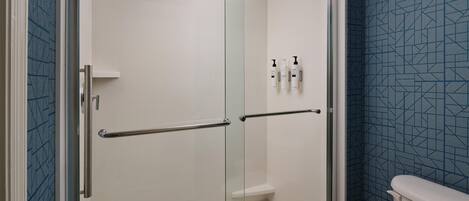 A glass-enclosed shower stall with a white tiled wall, a metal handrail, and a toilet with a blue patterned wall in the adjacent room.