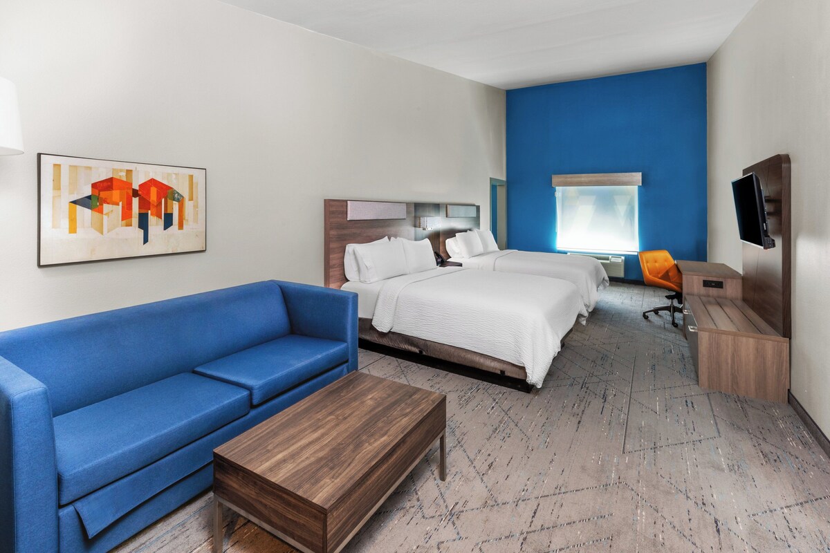Habitación de hotel con un sofá azul, dos camas, un escritorio de madera, un televisor y un cuadro en la pared.