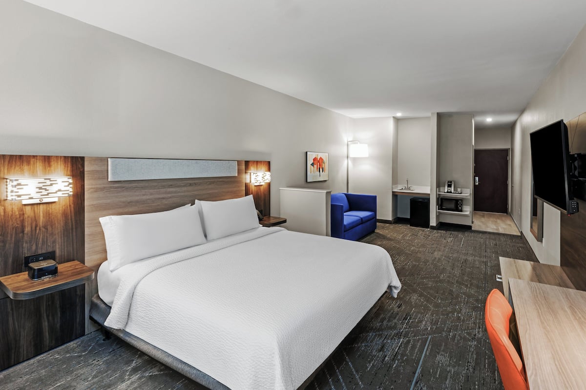 Ein Hotelzimmer mit einem großen Bett, einem Schreibtisch, einem Stuhl, einem Fernseher und einer kleinen Küchenzeile.